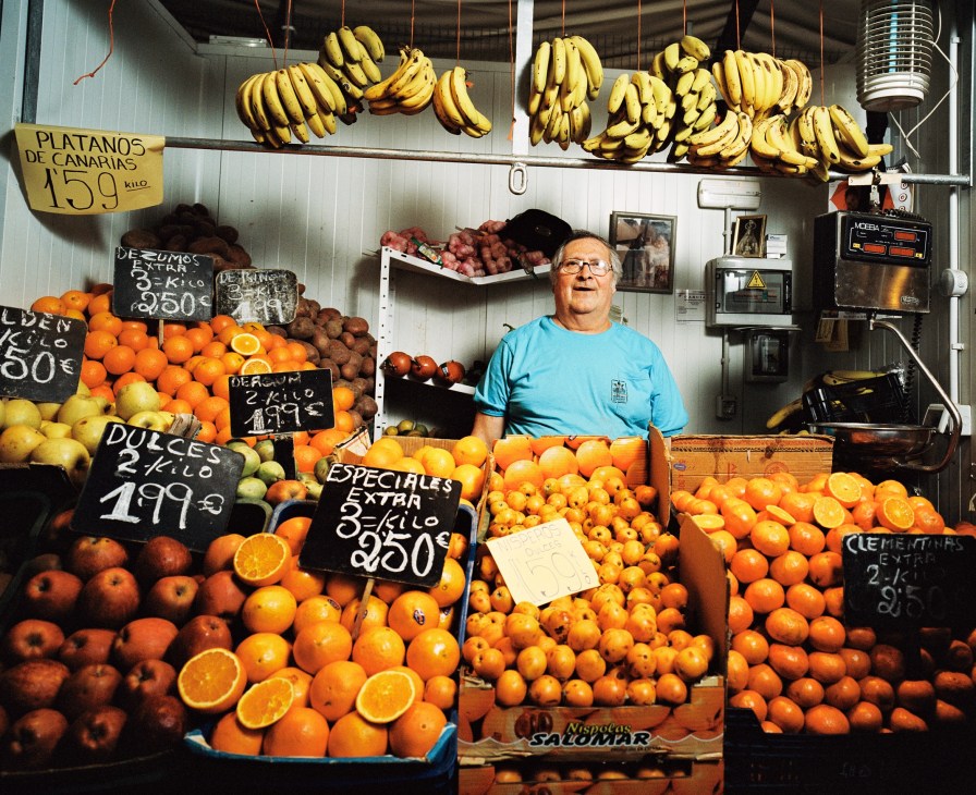 Fruit market stallholder in Cádiz surrounded by oranges, bananas, and other produce with handwritten price signs.