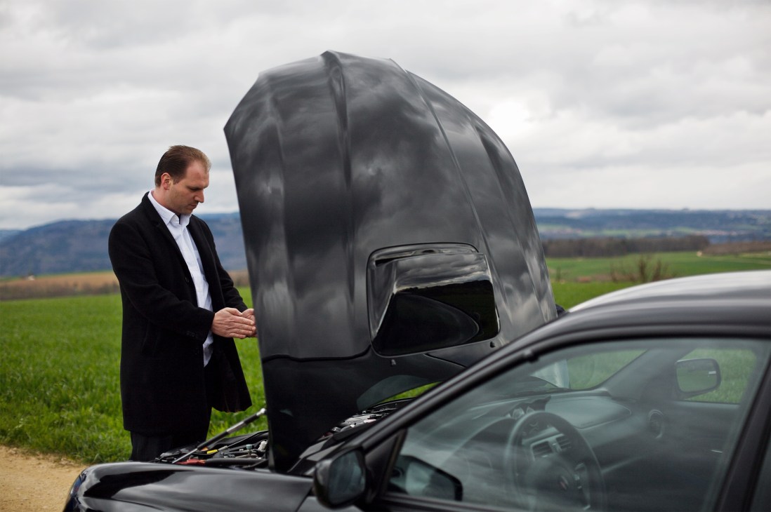 Man in blazer stands beside a Subaru with its hood up in a rural landscape.