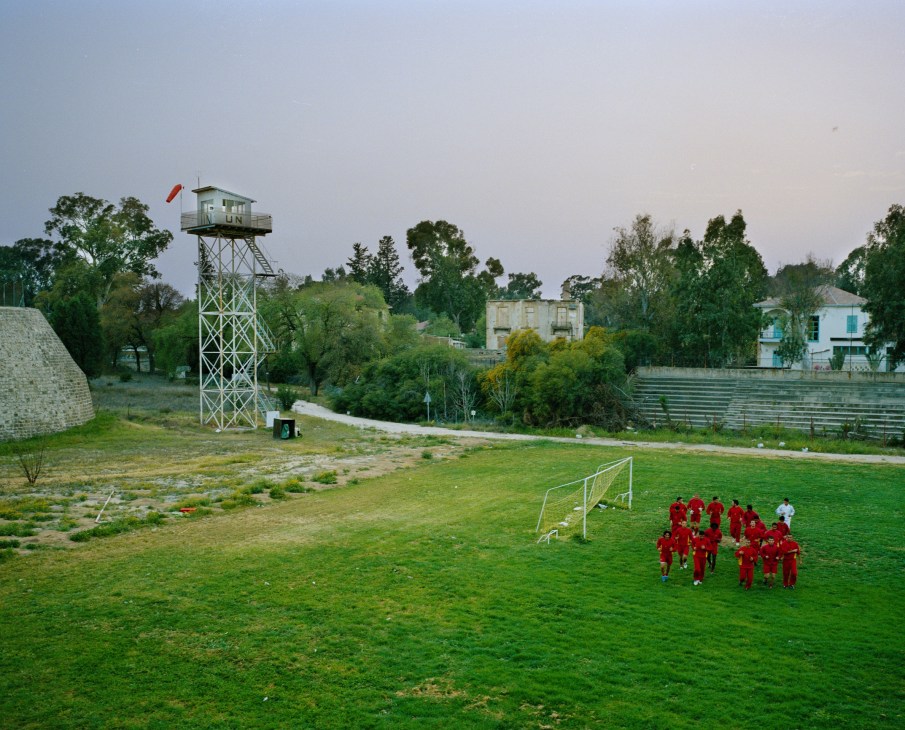 A disused UN watchtower with a Turkish flag overlooks a football pitch where a team in red jerseys trains in Northern Cyprus.