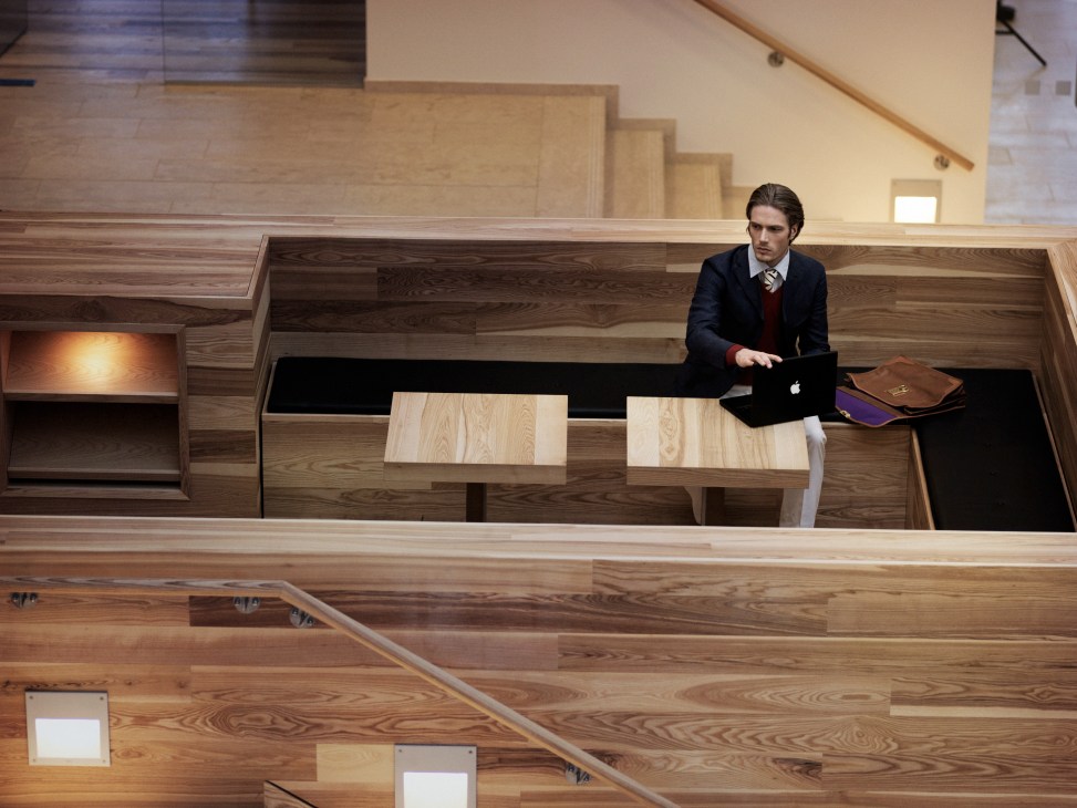 Man in navy blazer and tie working at wooden desk in modern office with ash wood booth seating.