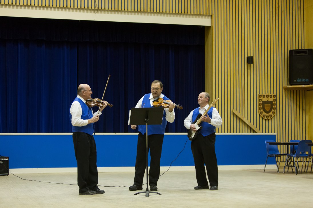 Three men in blue vests and white shirts play violins on stage with blue curtains behind them.