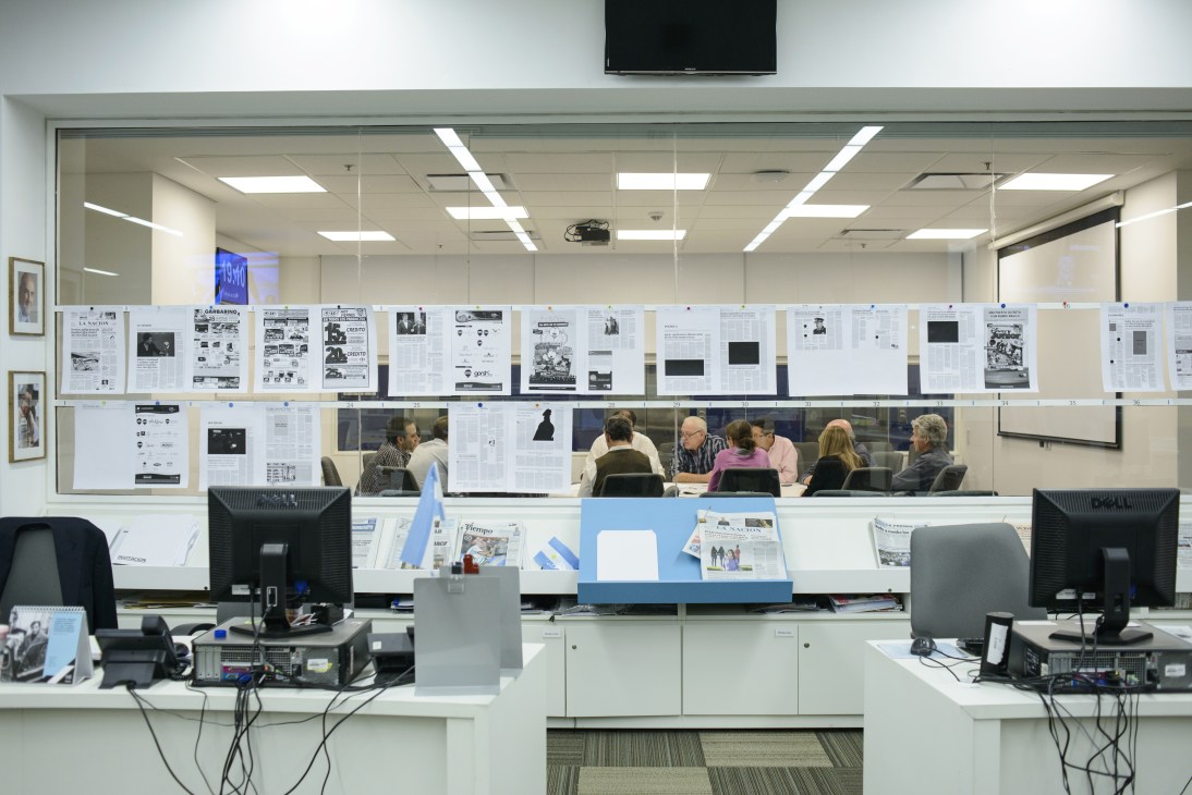 La Nación newsroom with pages pinned to glass wall behind staff meeting at tables.
