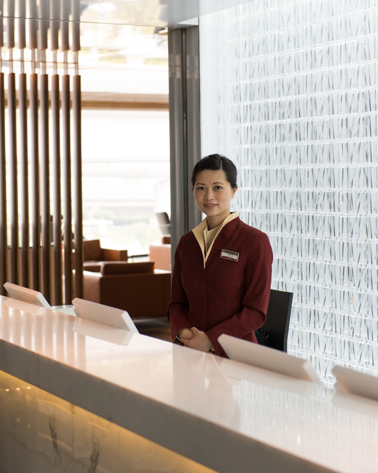 Hotel concierge staff member in burgundy uniform stands at modern reception desk.