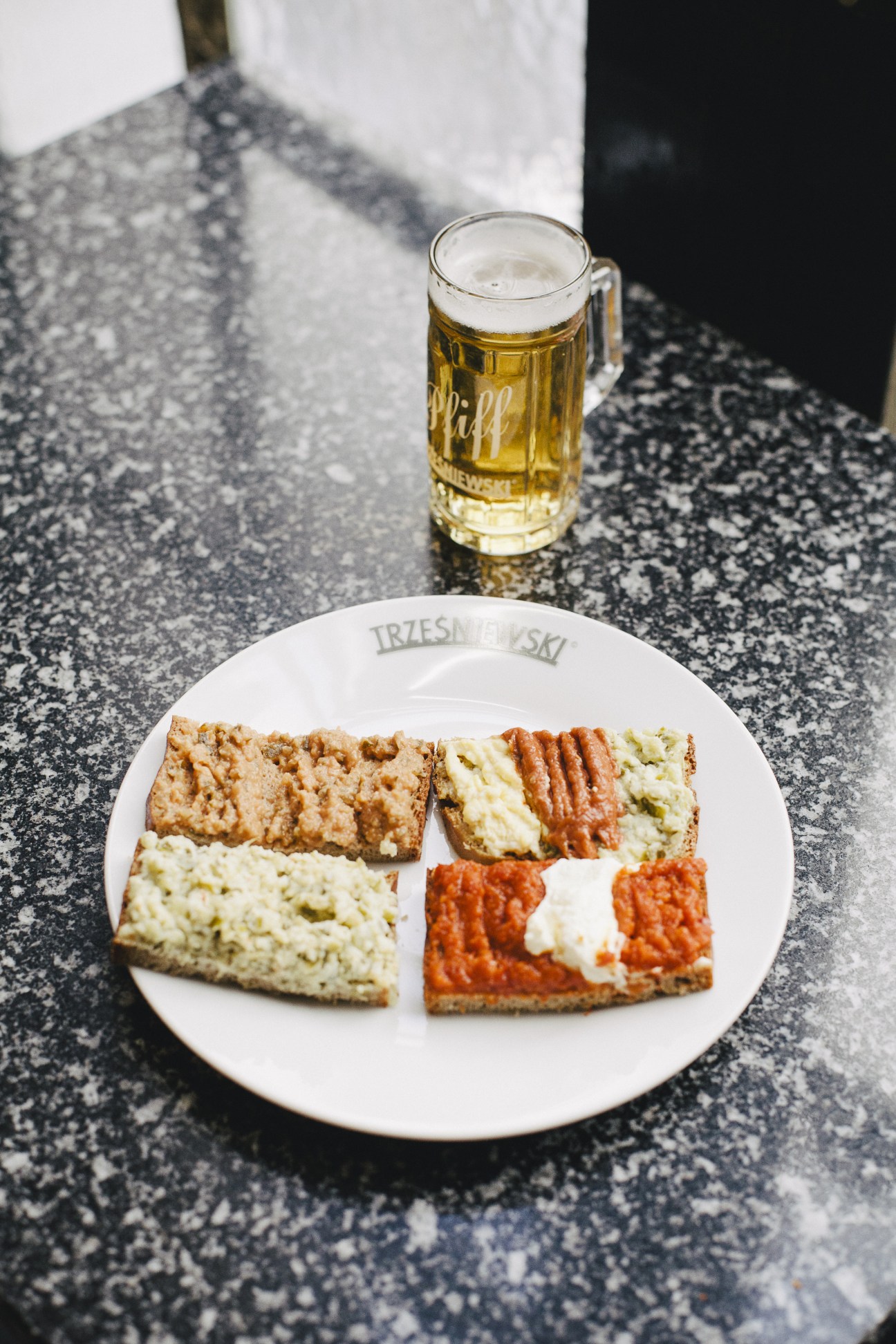 Open-faced sandwiches on a Trzesniewski plate with a glass of beer on a speckled table.