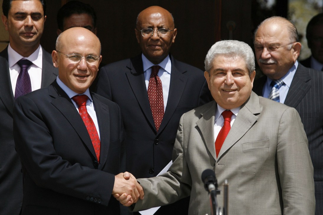 Two men in business suits shake hands while standing with other officials at a microphone.