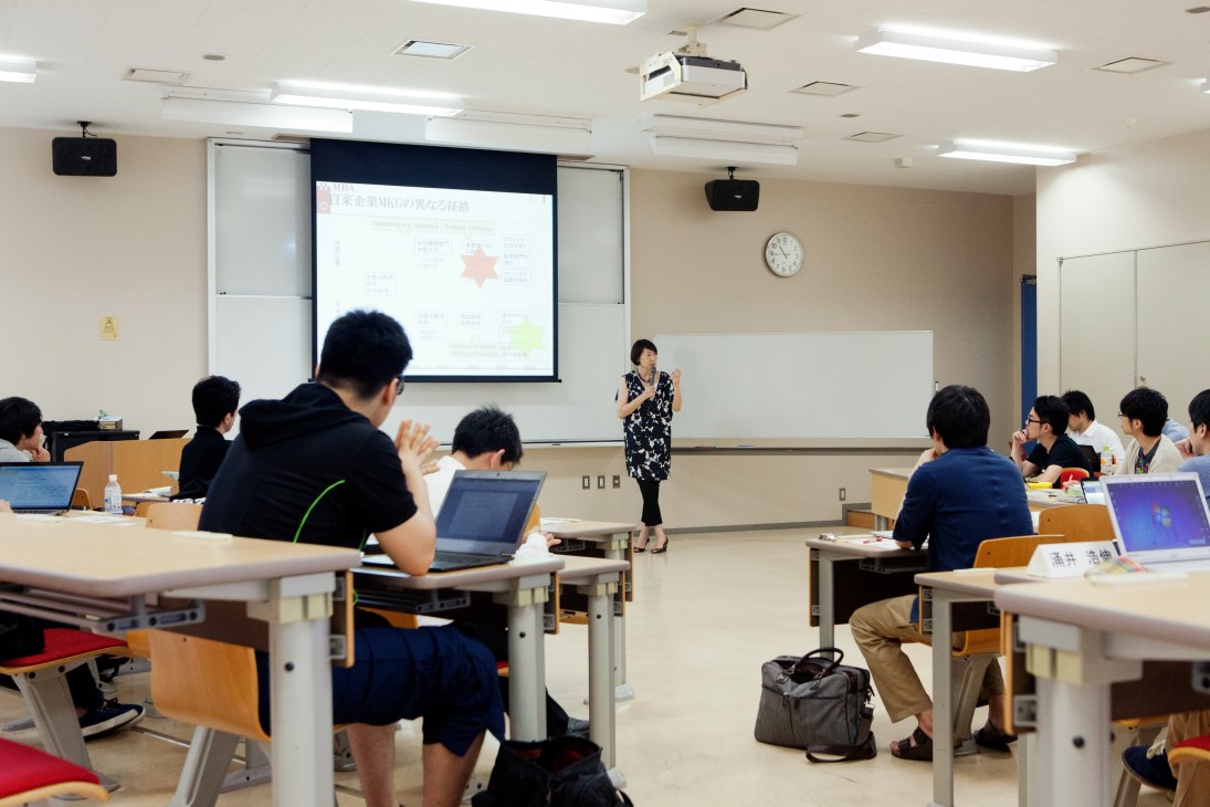 MBA students seated at desks with laptops listen to a female professor presenting a business strategy diagram on a projection screen.