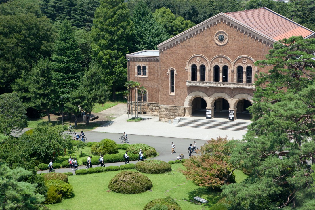 Aerial view of a brick building with arched windows on Hitotsubashi University campus surrounded by landscaped gardens and students walking on paths.