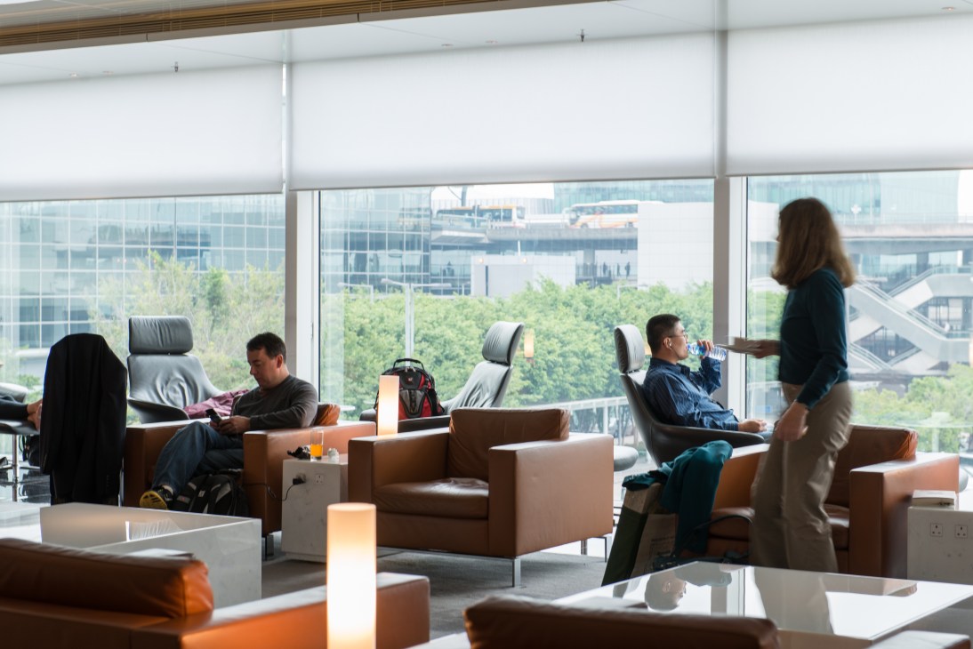 Modern airport lounge with brown leather seating, floor-to-floor windows overlooking cityscape and green trees, passengers relaxing and working.