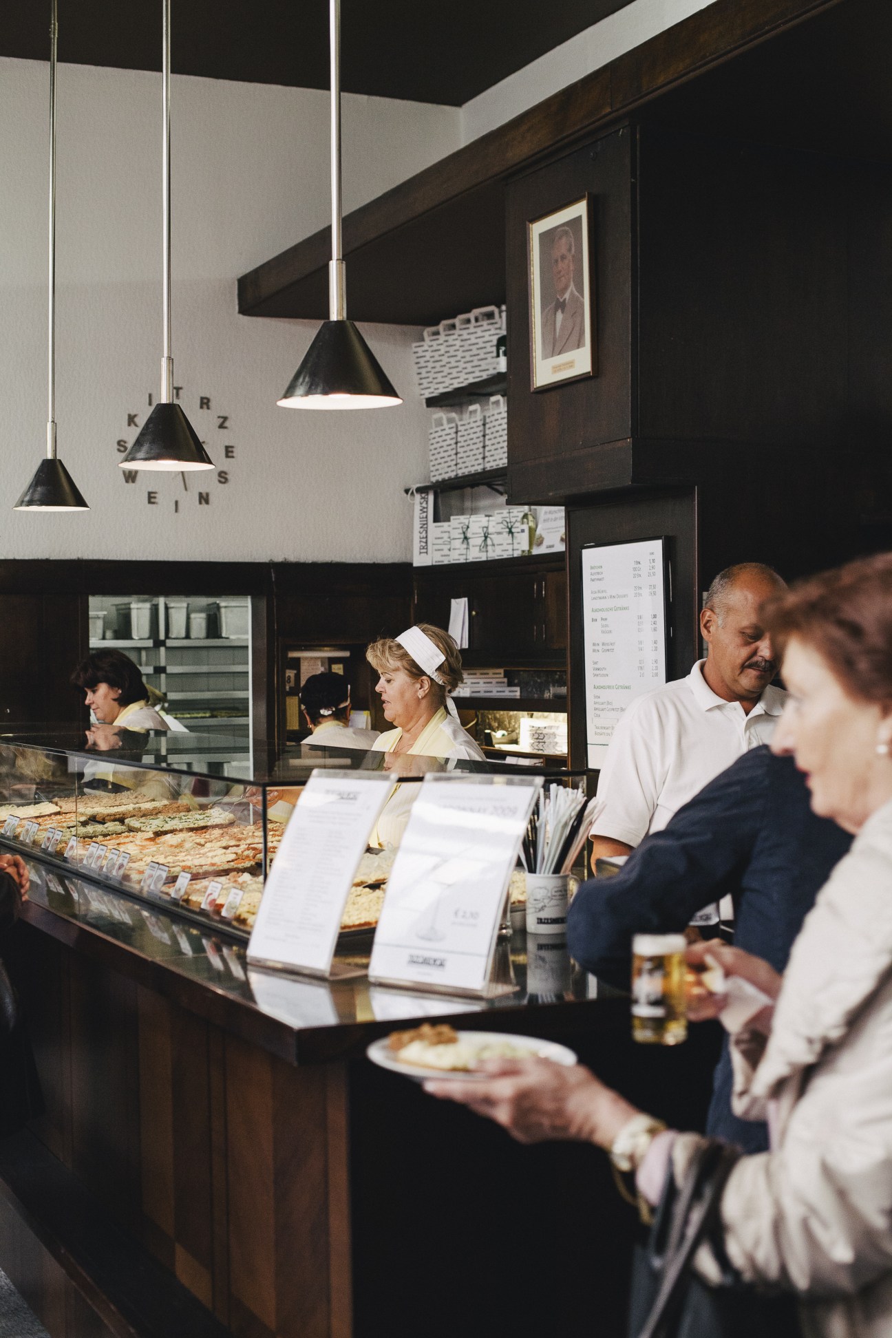 A Viennese bakery counter with staff serving customers, displaying pastries and sandwiches under pendant lights.
