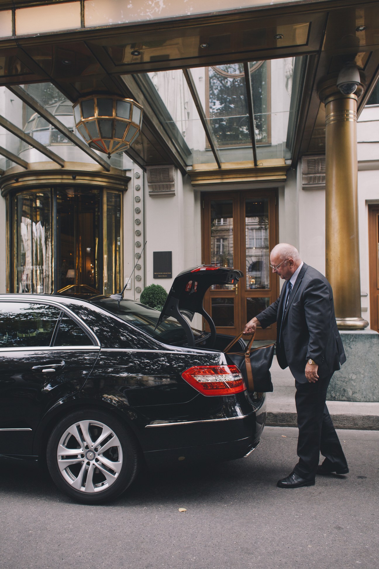 An elderly chauffeur in a dark suit opens the trunk of a black Mercedes car outside a Vienna hotel entrance.