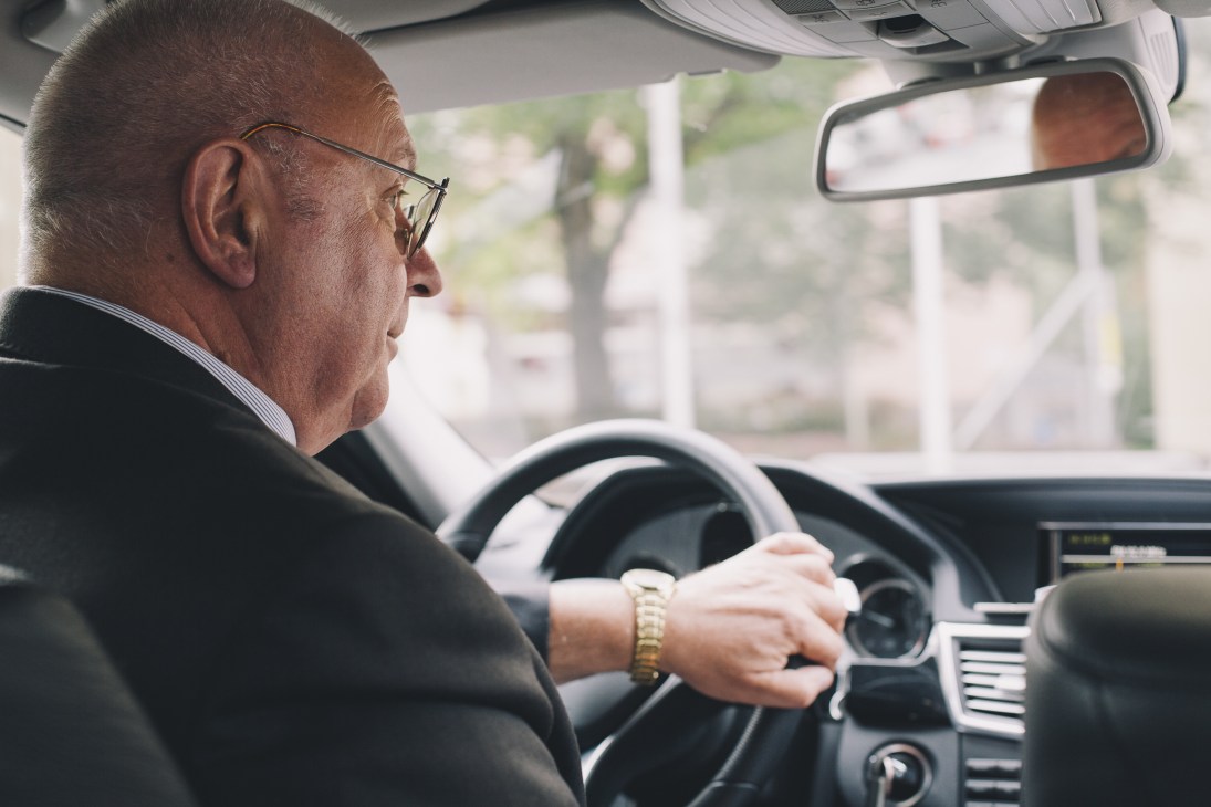 Older man wearing glasses driving a luxury car, viewed from the side in the driver's seat.