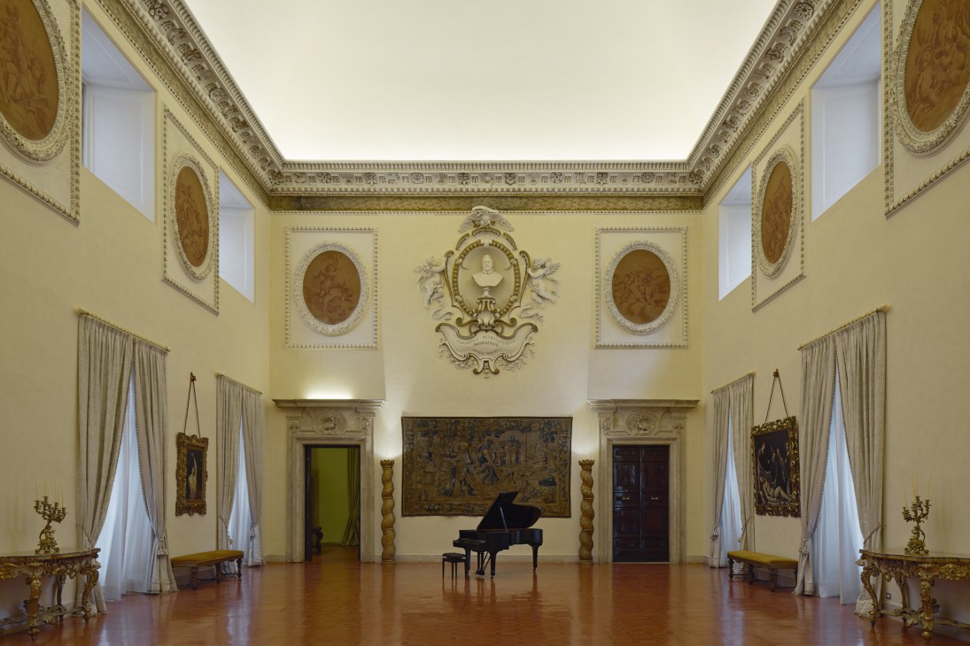 Grand palatial reception room with ornate cream and gold ceiling, portrait medallions, grand piano, and classical architectural details.