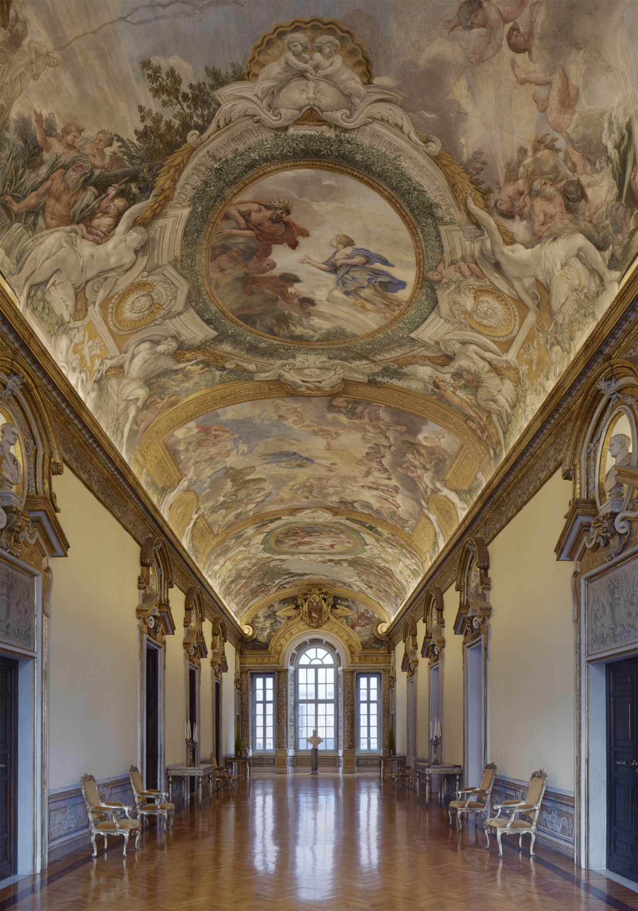 A grand palazzo gallery with ornate frescoed ceiling, gold-trimmed architectural details, and tall windows at the far end.