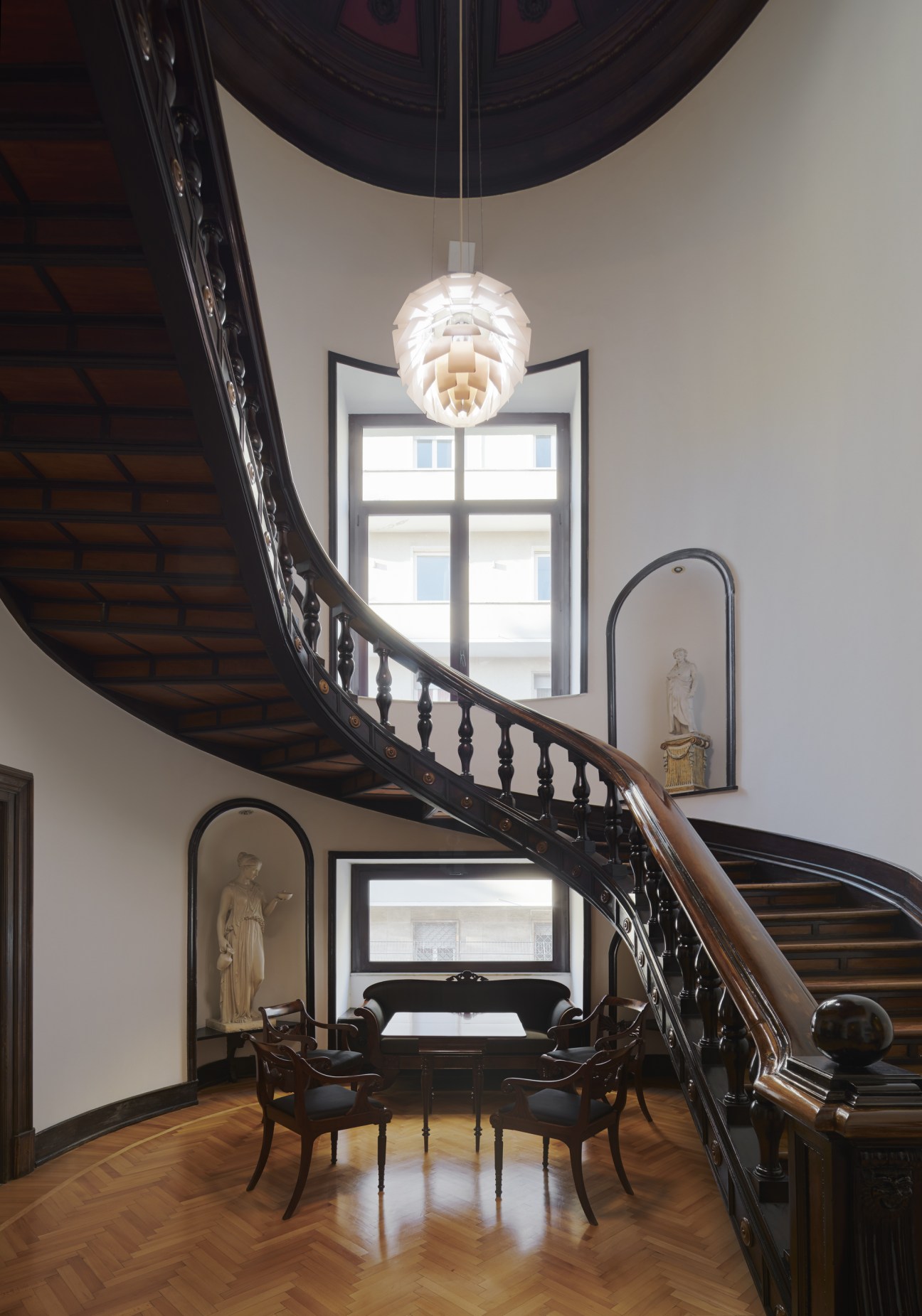 Grand wooden staircase with ornate black railings, sculptural niches, and modern pendant light in Danish embassy.