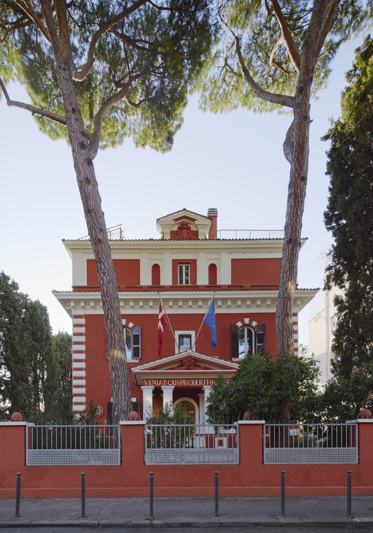 Red and white striped Italian Renaissance building with tall pine trees and diplomatic flags flying from the facade.