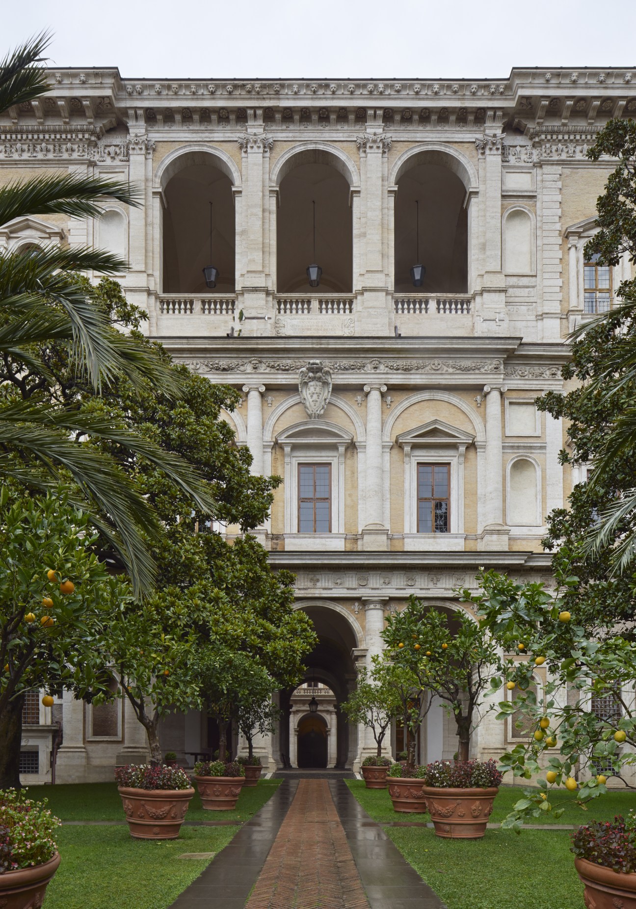Classical palazzo facade with arched openings viewed from a formal garden with citrus trees and potted plants.
