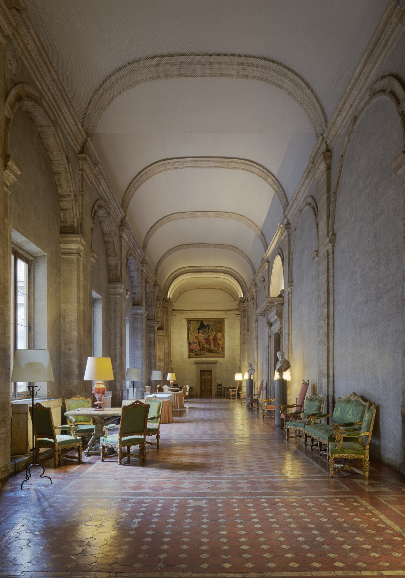 A long classical corridor with barrel-vaulted ceiling, ornate columns, period furniture, and paintings in an embassy palace.