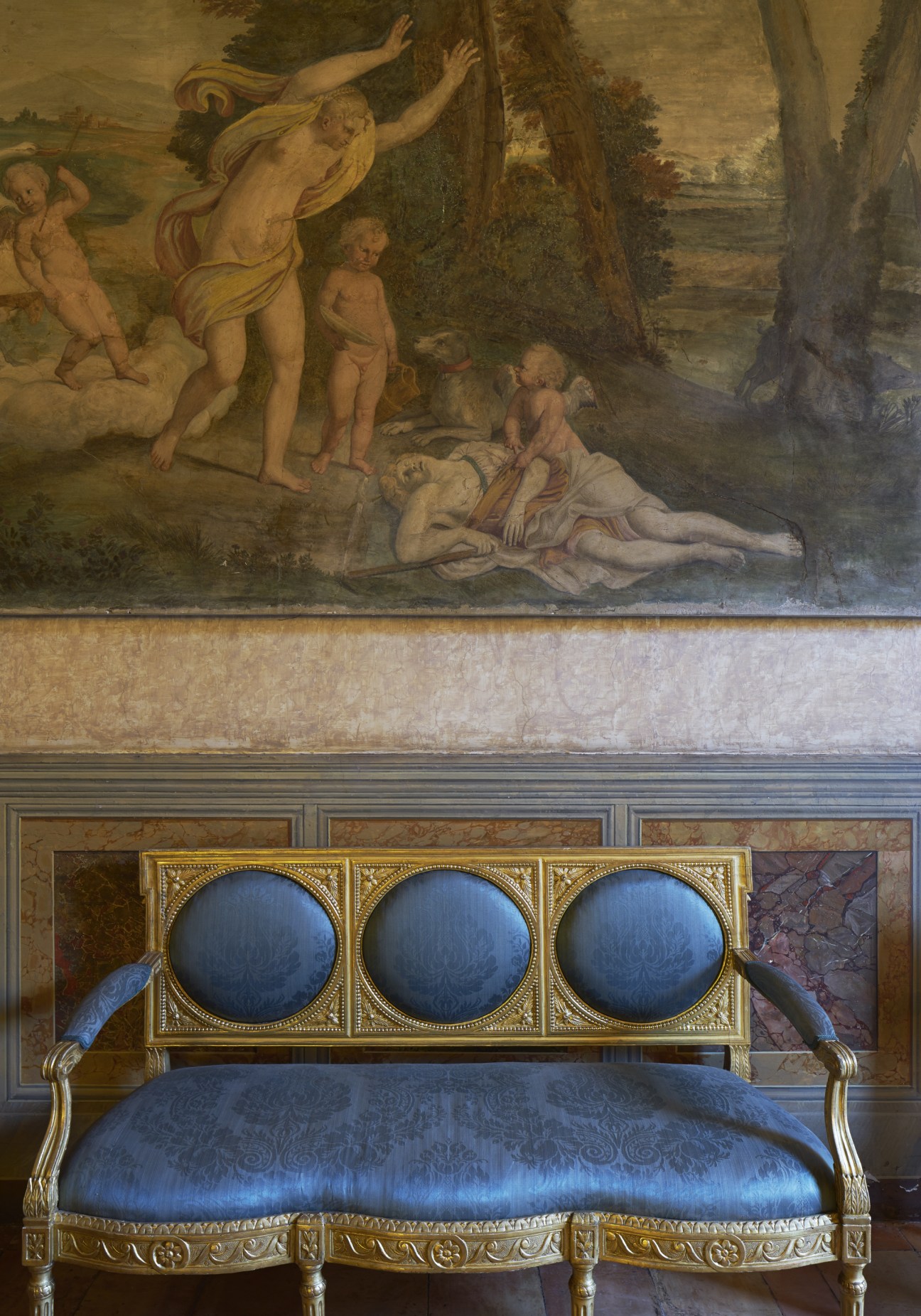 Renaissance-era chair with blue silk upholstery and ornate gold frame beneath a classical fresco depicting mythological figures.