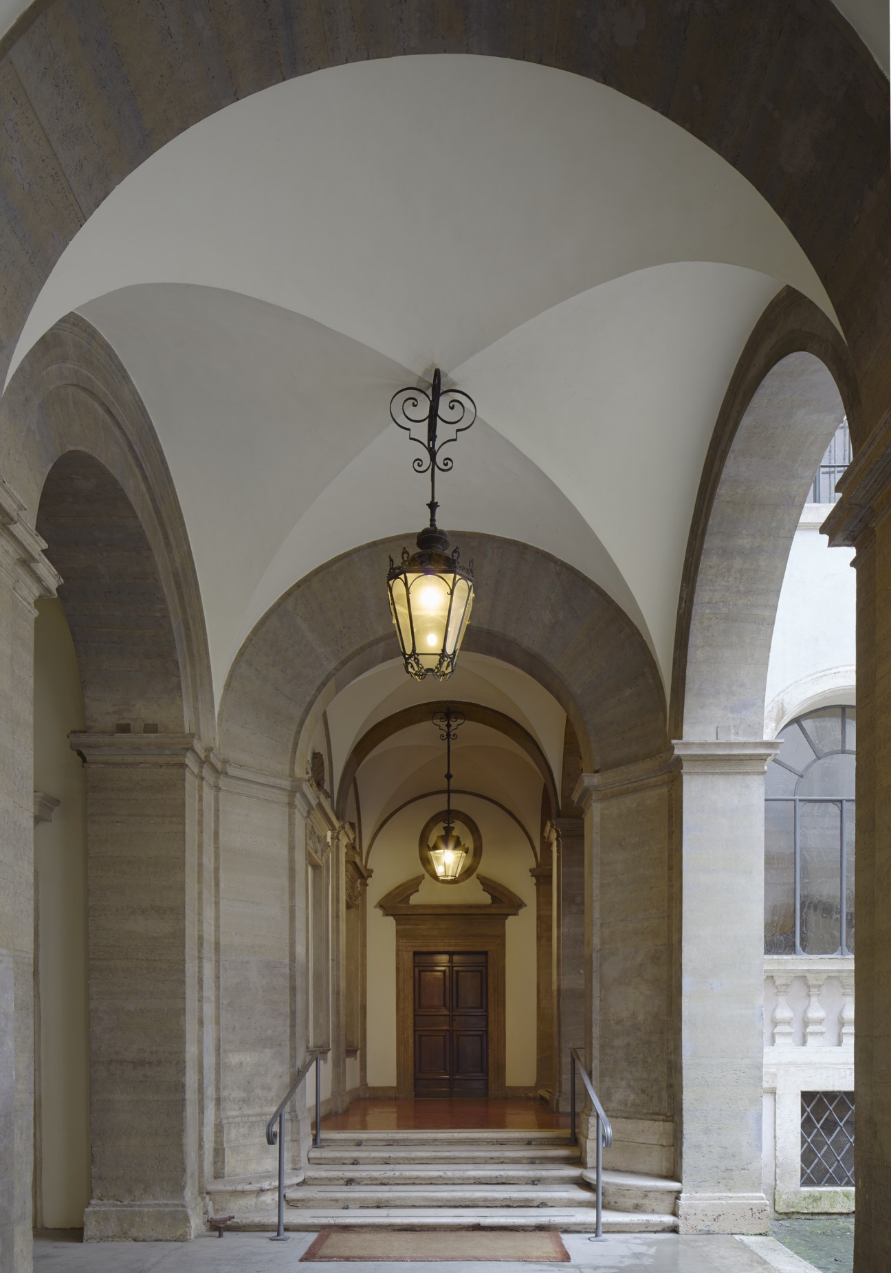 Vaulted Renaissance arcade with stone columns, hanging lanterns, and a wooden door at the end of a palazzo courtyard.