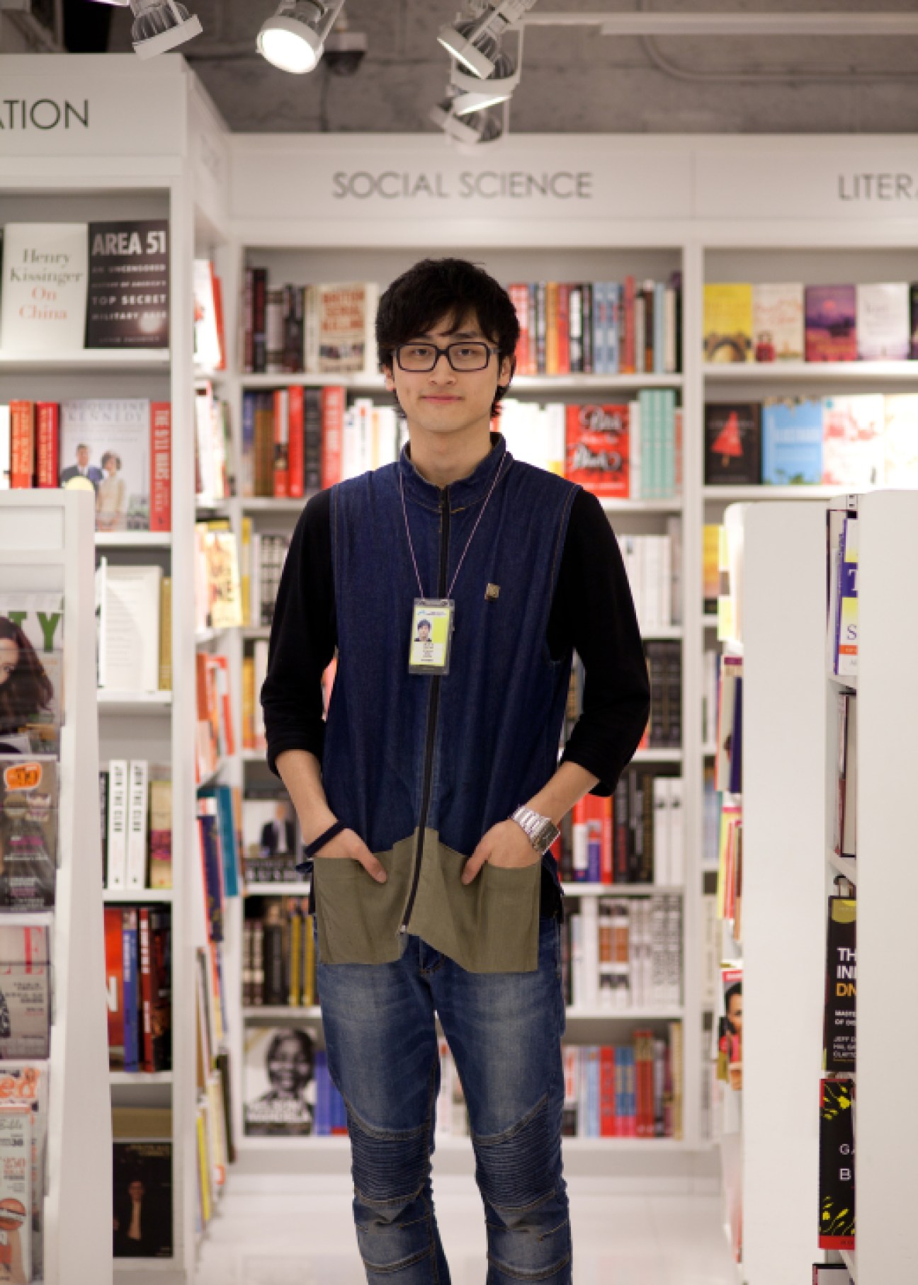 Young man wearing glasses and navy vest stands in front of well-stocked bookshelves at Page One airport kiosk.