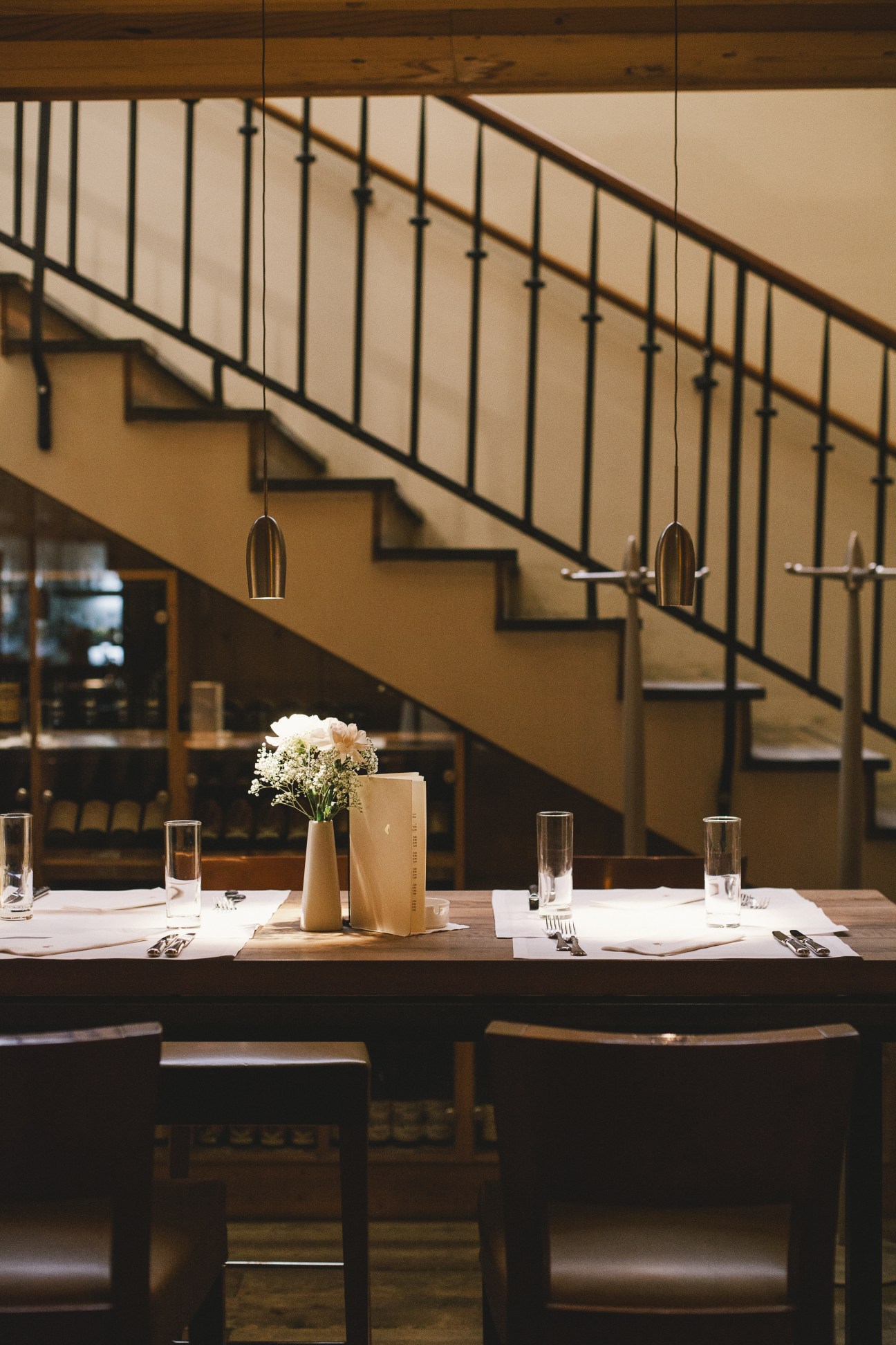 A minimalist dining table set with white napkins, glasses, and a white flower arrangement under a modern staircase.