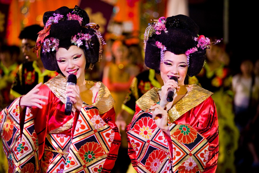 Two performers in elaborate red and gold costumes with pink flowers in their hair sing into microphones during a getai performance.