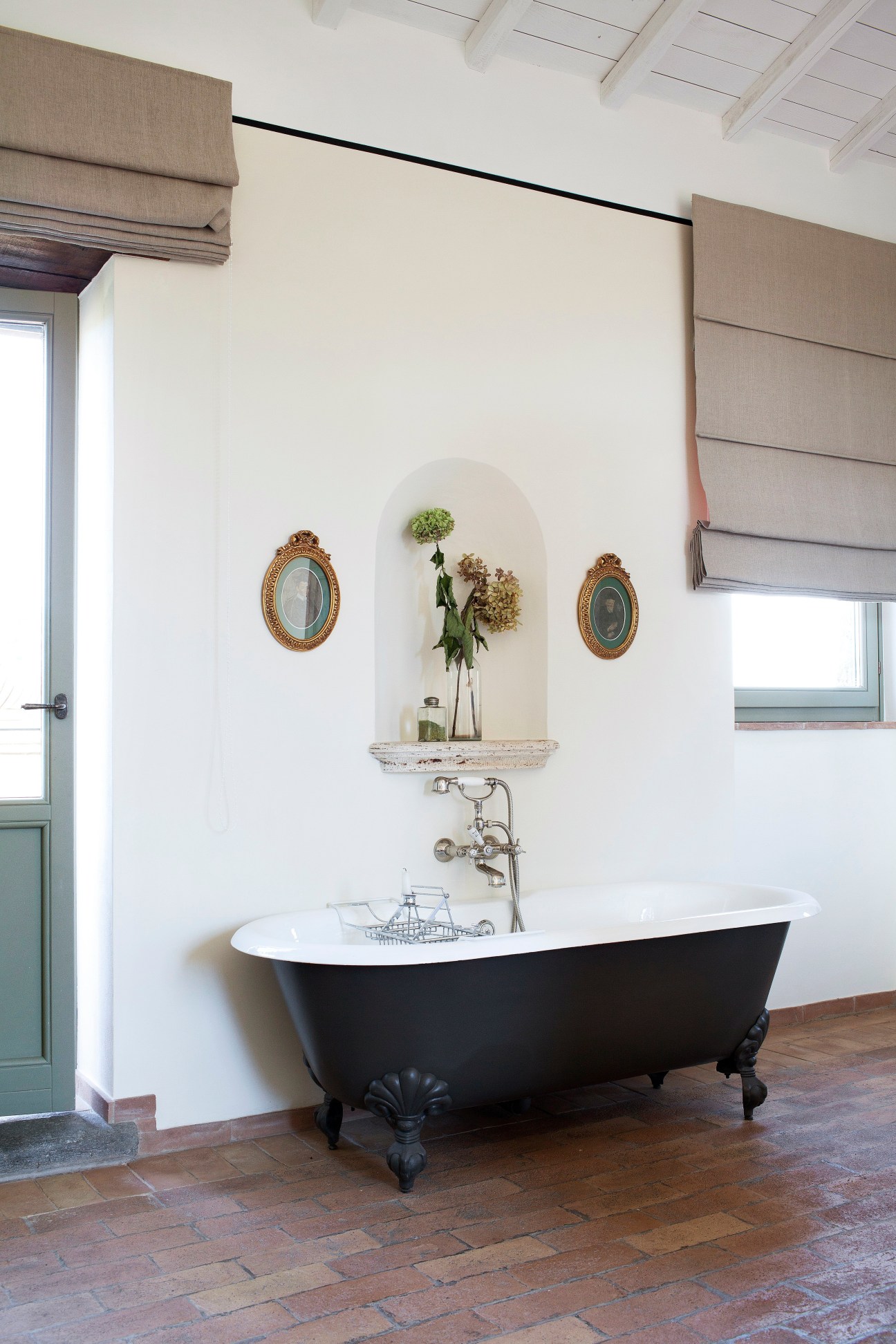 Black clawfoot bathtub with white interior against white wall with arched niche holding flowers and framed portraits.