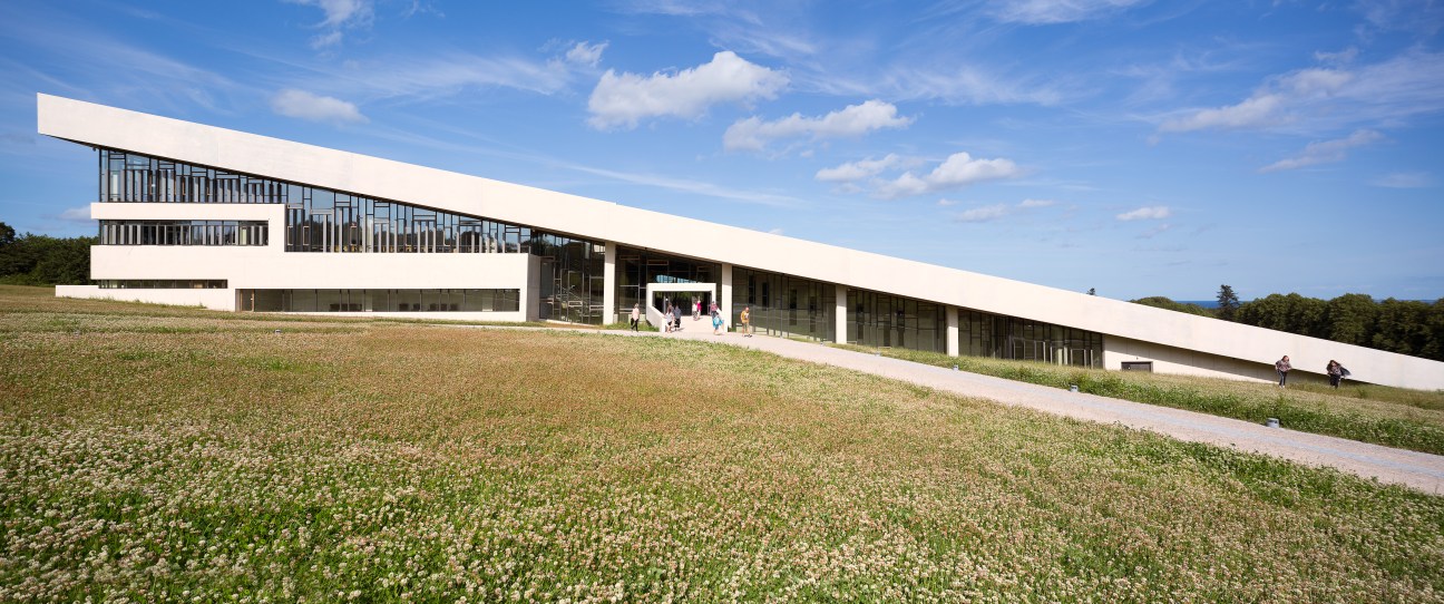 Moesgaard Museum's modern white building with sloped roof sits on a grassy hillside under blue sky.