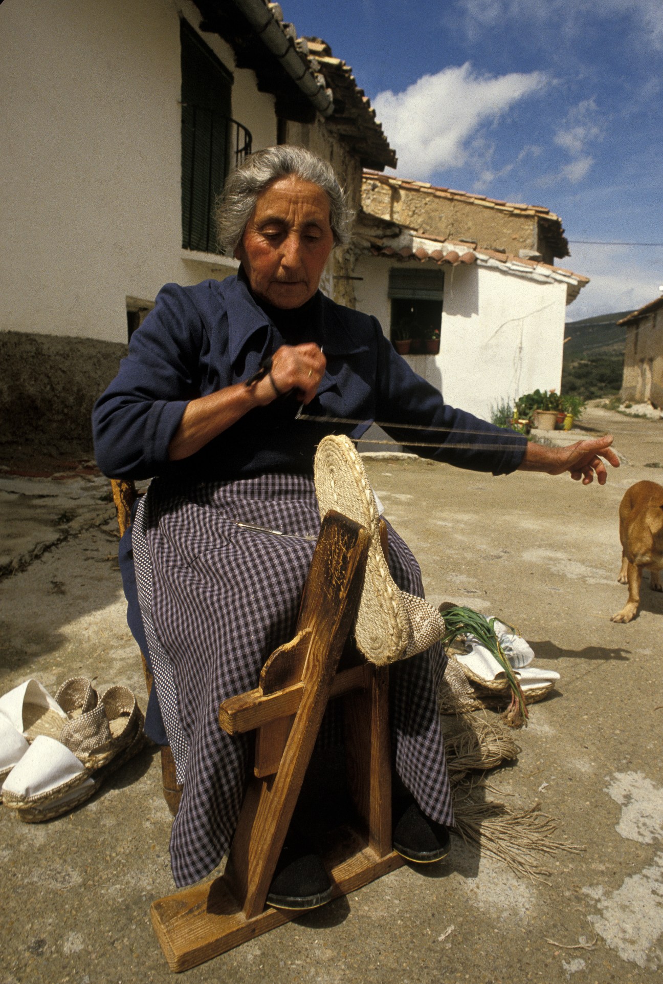 Elderly woman in blue jacket demonstrating traditional flip-flop weaving on wooden loom outside rural home.