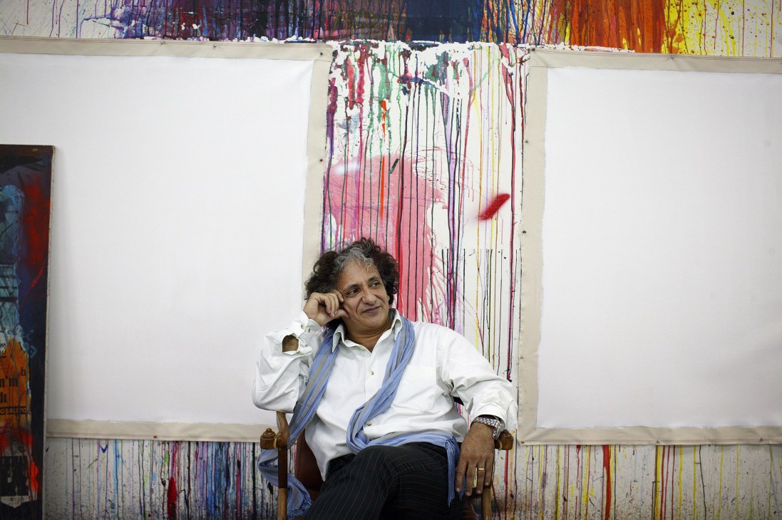 Artist Bedri Baykam sits in his Istanbul gallery studio, positioned in front of a colorful abstract painting with vertical drips and streaks.