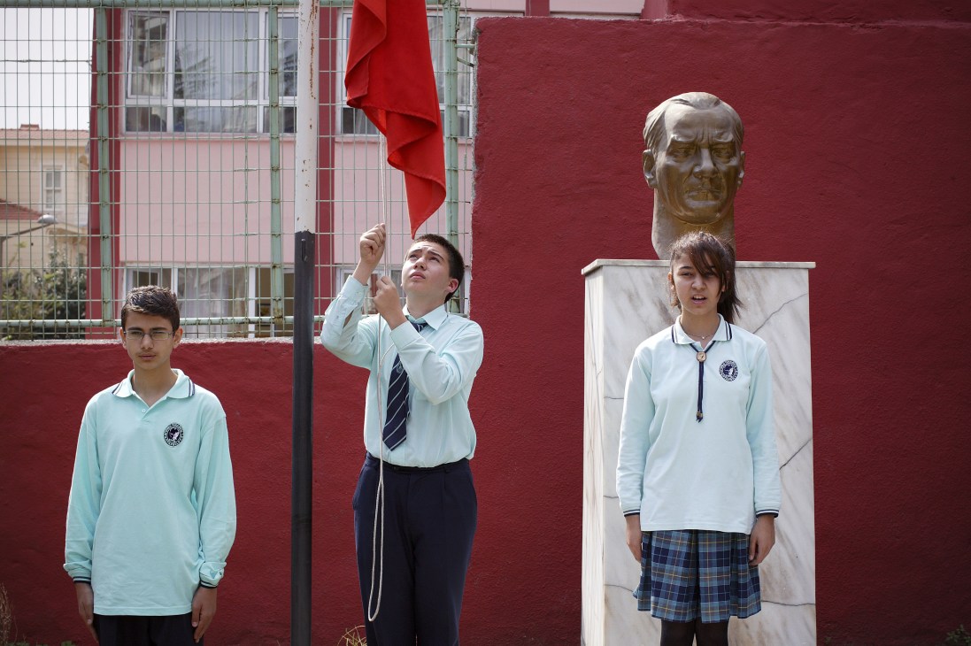 Three students in school uniforms stand in a playground near a bronze bust of Atatürk mounted on a white pedestal against a red wall.