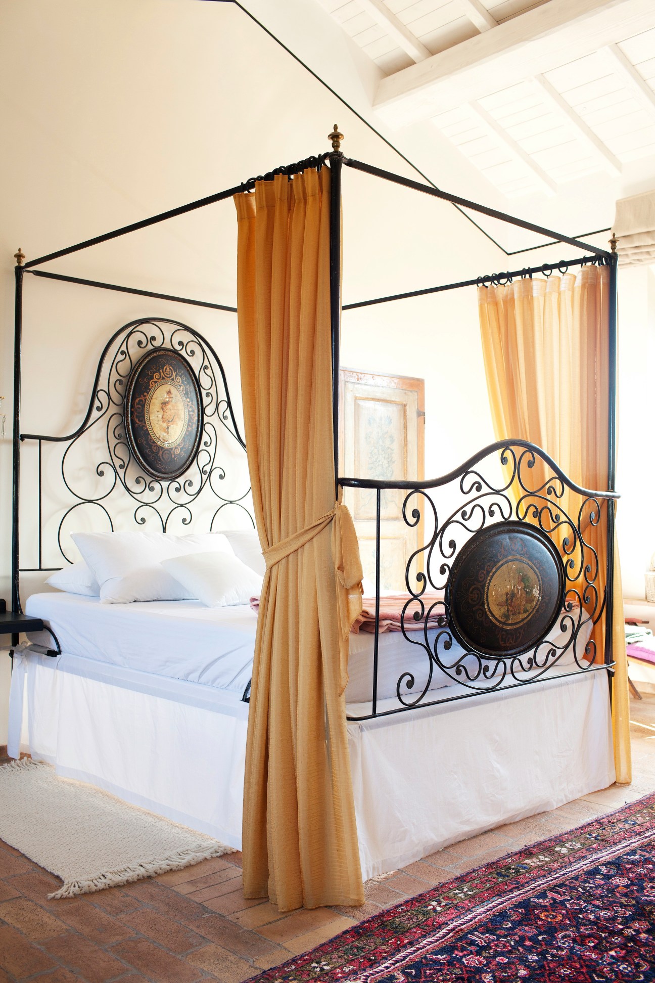 Medieval bedroom with ornate iron four-poster bed, gold curtains, and decorative wall medallion at Corte della Maestà in Italy.
