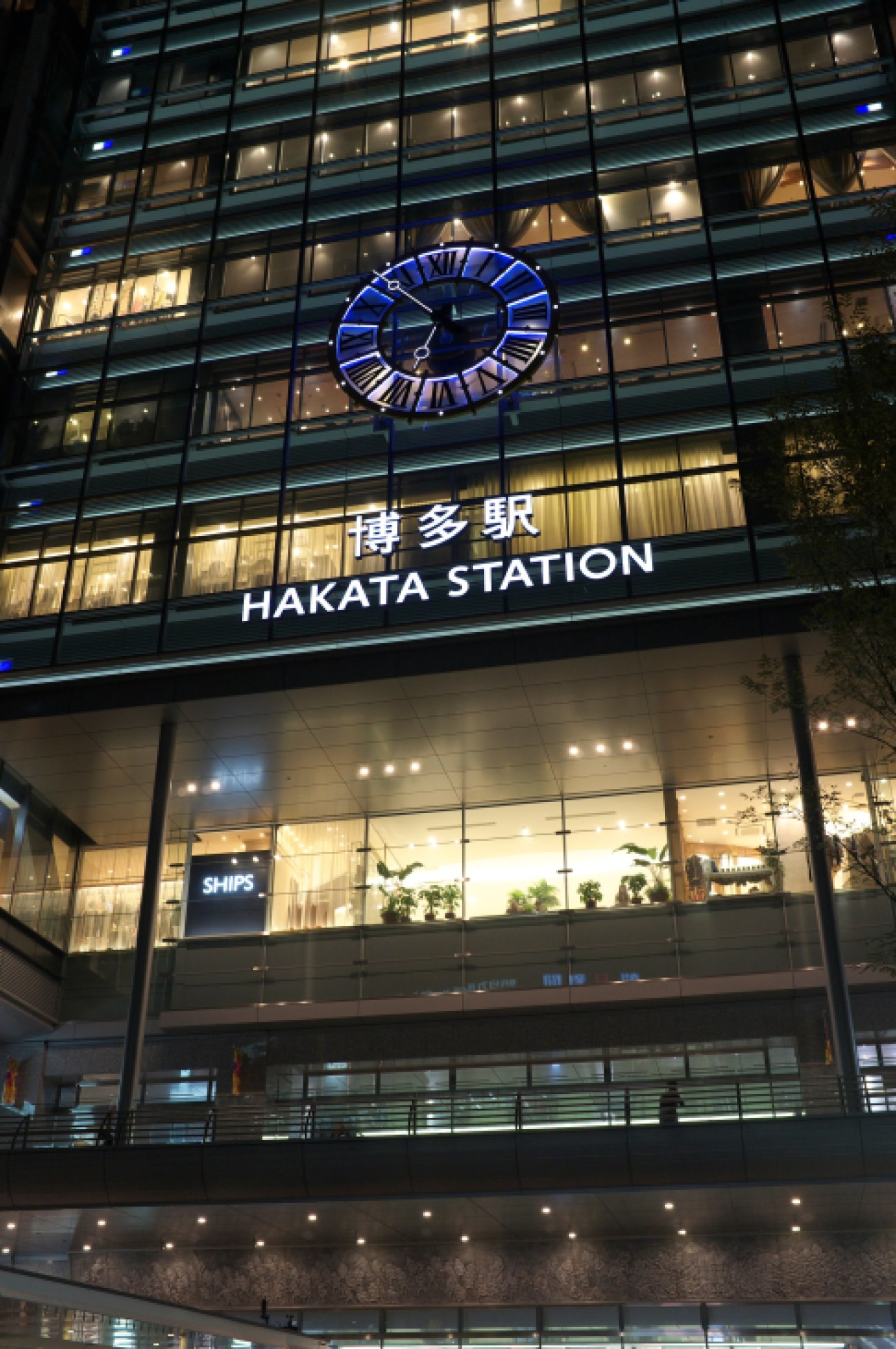 Hakata Station's modern facade at night with illuminated blue clock and Japanese signage.