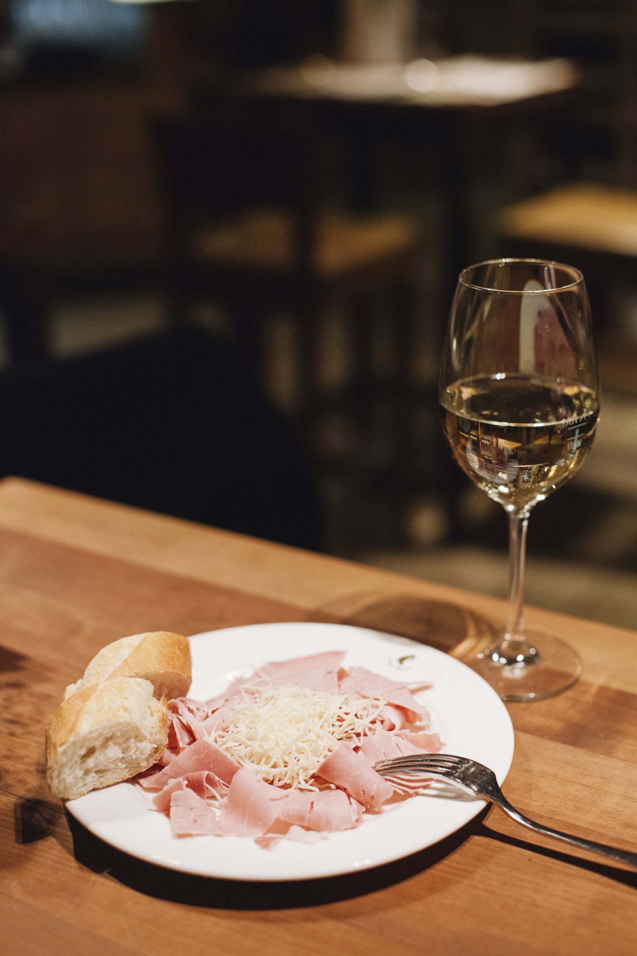 A glass of white wine and a plate of sliced ham with shredded cheese and bread on a wooden table.