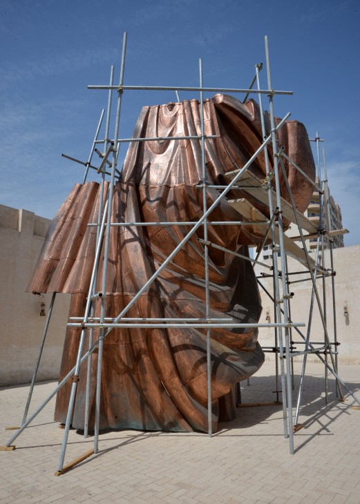 Large bronze sculptural head under construction with metal scaffolding, displayed outdoors in bright sunlight.