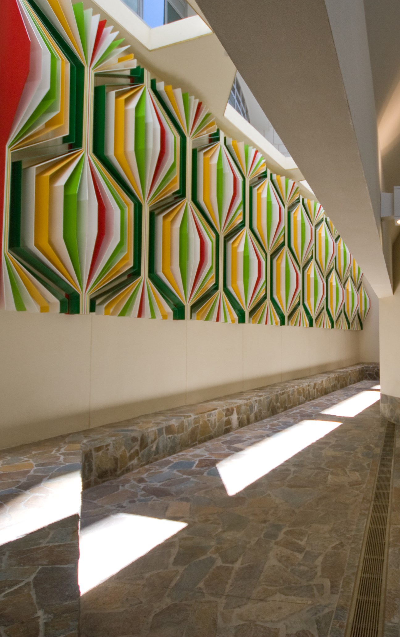 Colorful geometric artwork with radiating striped panels in red, yellow, green and black inside a modern corridor with stone flooring.