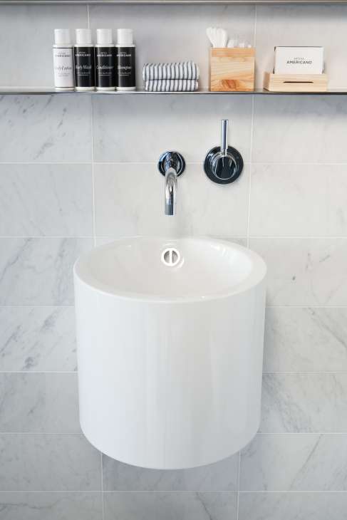 Hôtel Americano bathroom sink with marble tiles, chrome faucet, and branded amenities on shelf.