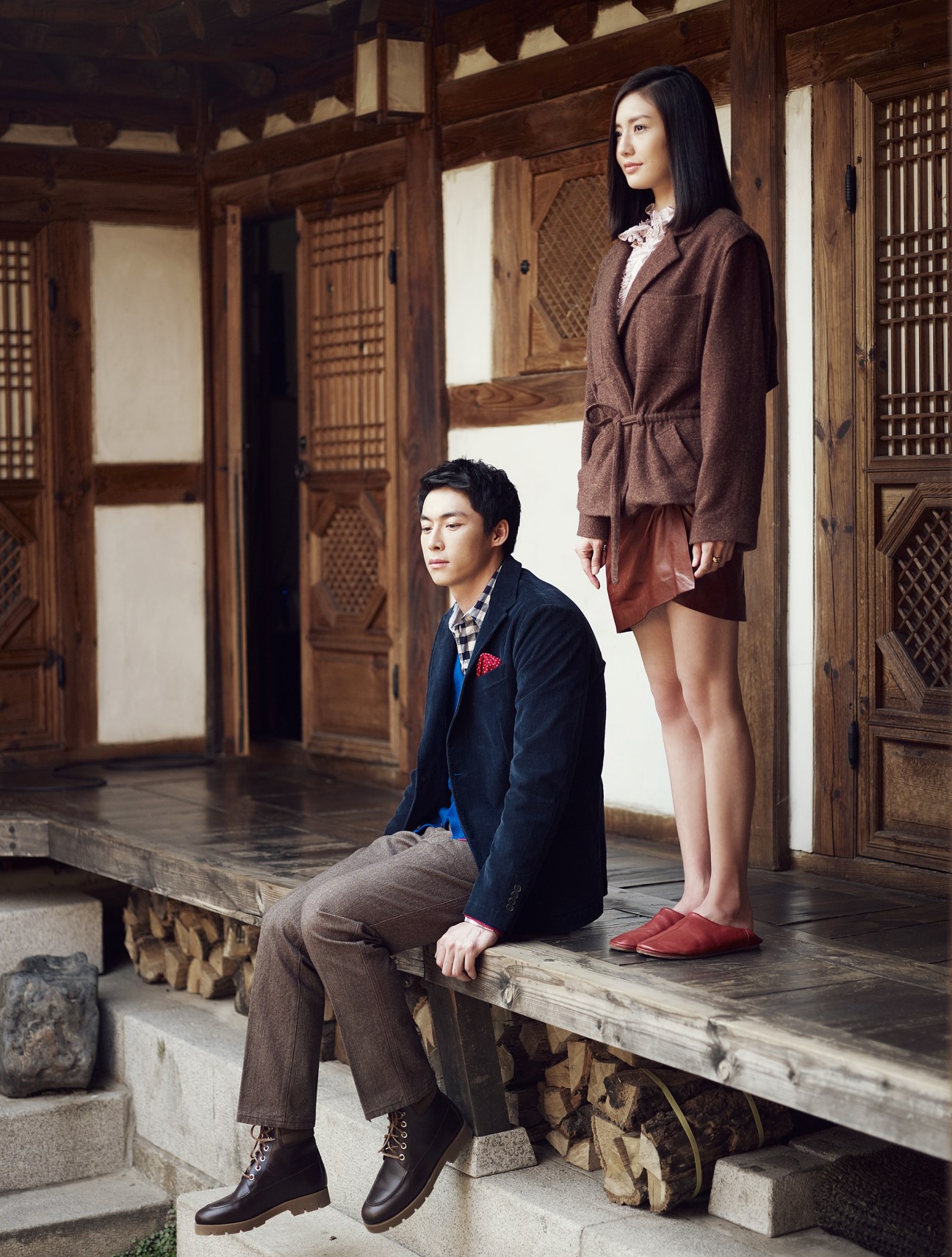 A young man sitting and a woman standing in a traditional Korean wooden hanok interior, wearing styled contemporary fashion.