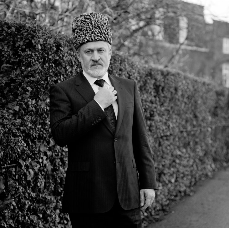 Akhmed Zakayev, Chechen resistance leader, in formal attire with traditional Caucasian papakha hat beside ivy-covered wall.