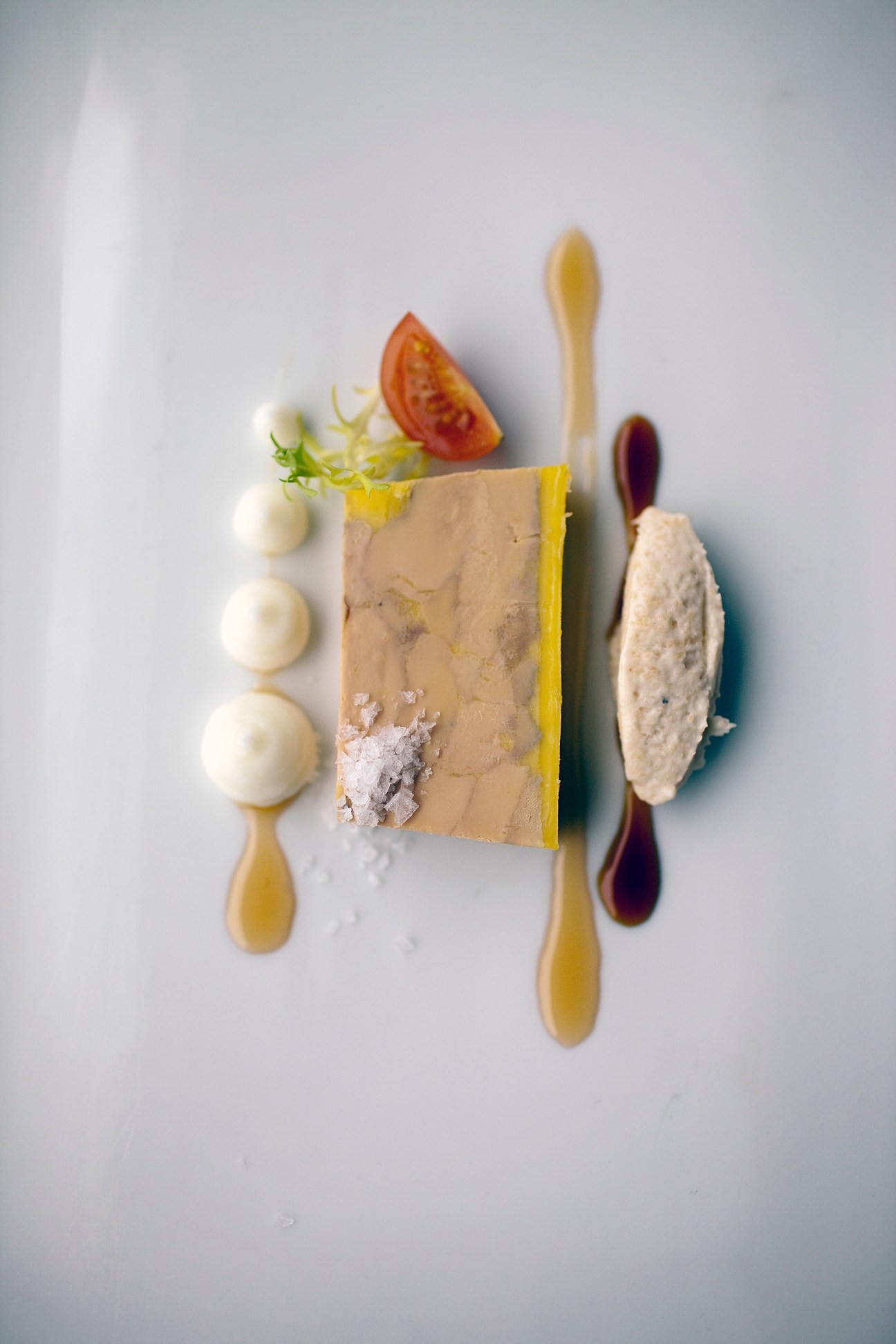 Plated foie gras dish with mango spheres, tomato, microgreens and sauce elements on white background.