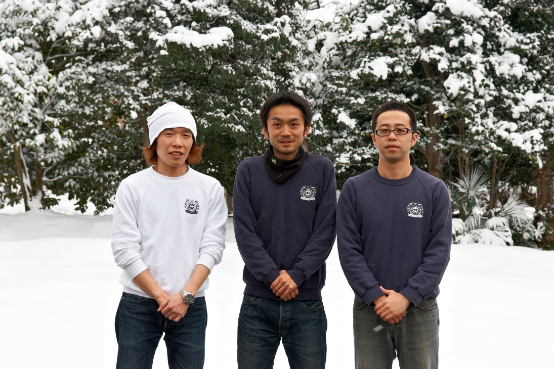 Three men wearing brewery-branded sweatshirts stand in snow with evergreen trees behind them.