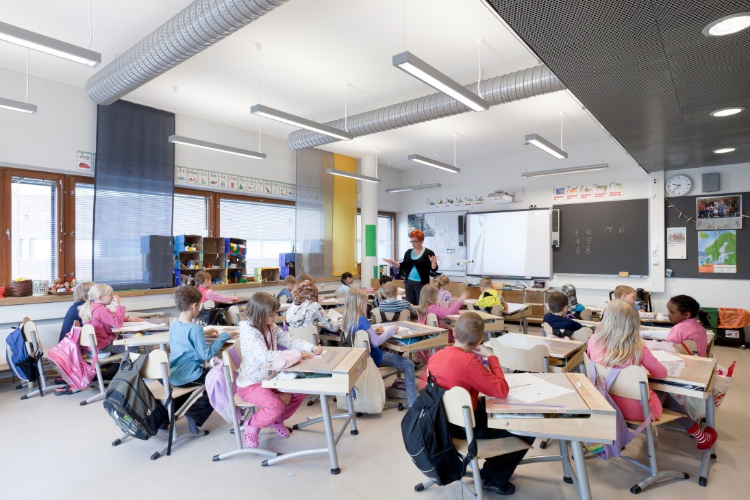 A bright classroom at Kirkkojärvi School with students seated at beech and steel desks, teacher instructing, and natural light from large windows.