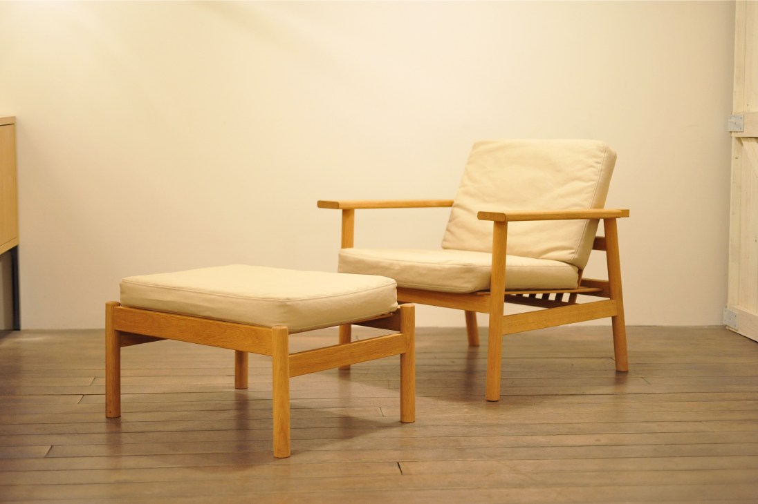Wooden armchair with cream cushions and matching ottoman in a minimalist studio setting.