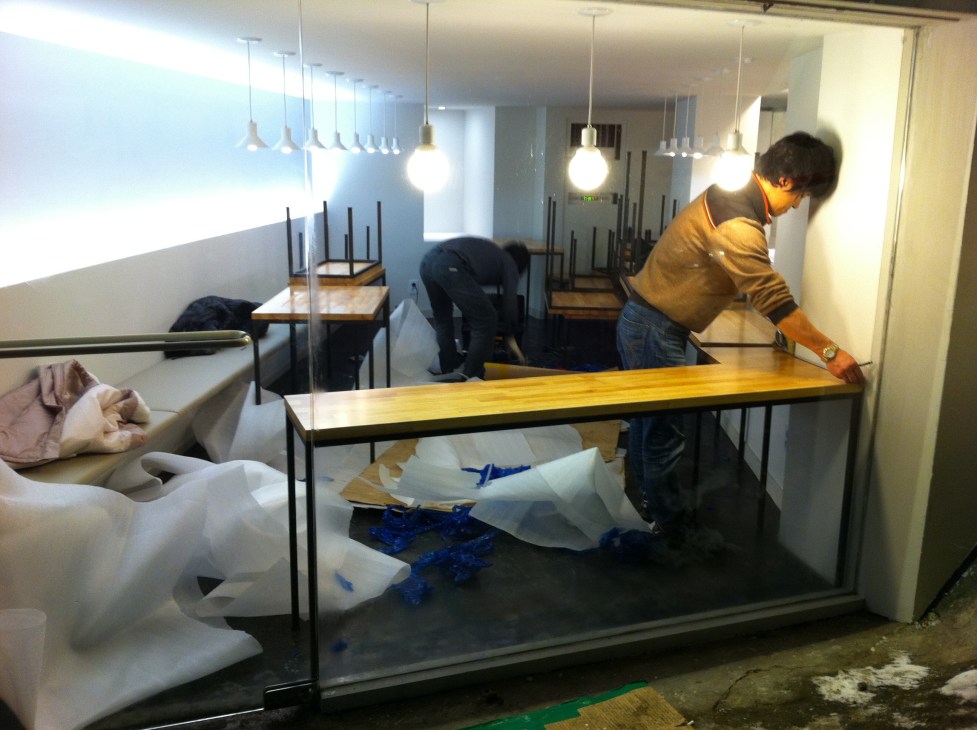 A storefront display under construction showing pendant lights and furniture pieces being installed.