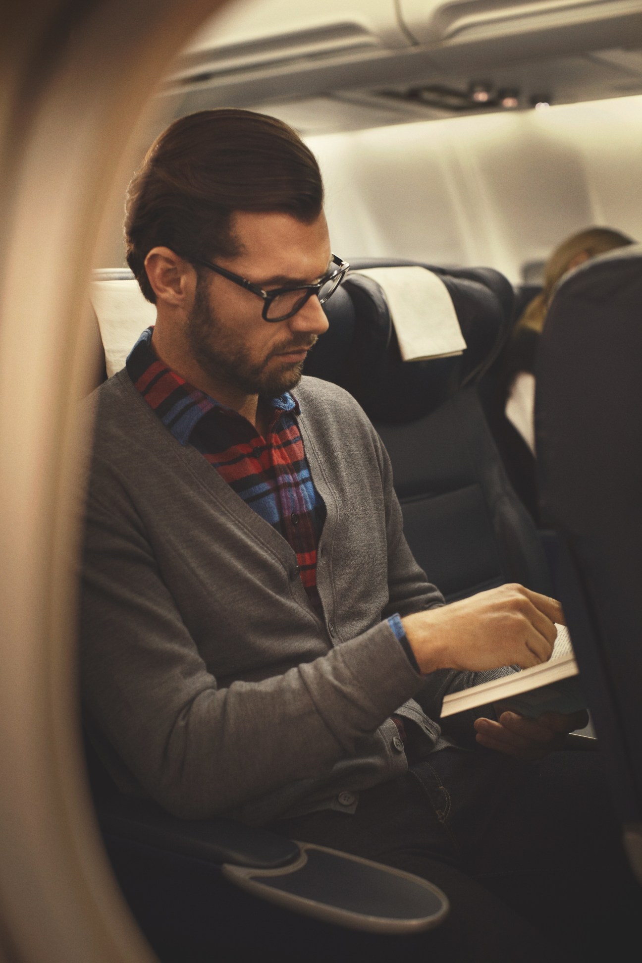 Man wearing Persol glasses and Zanone cardigan reading on an airplane.