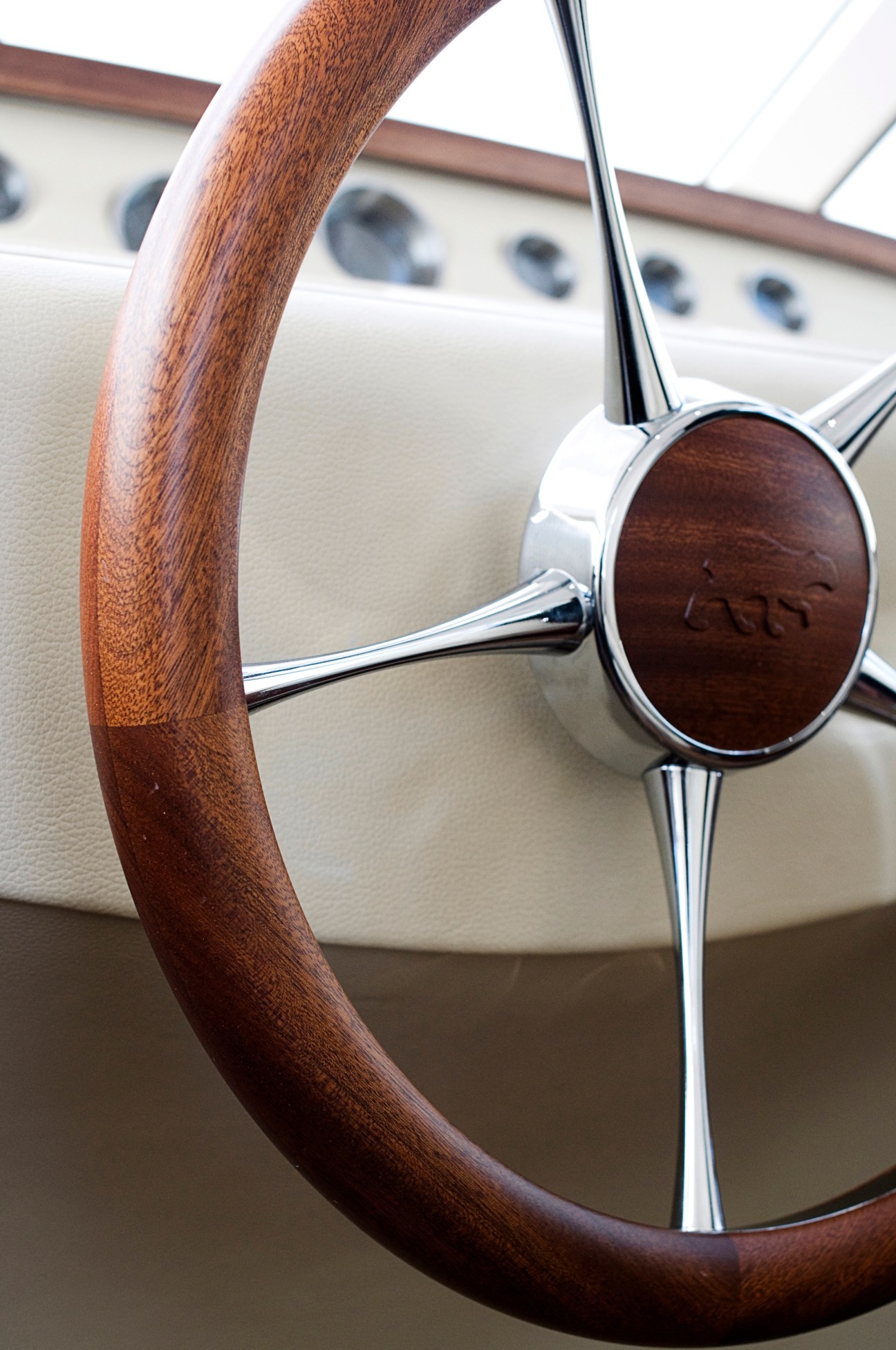 Close-up of Aguti yacht's wooden helm steering wheel with chrome details and cream-colored upholstery background.