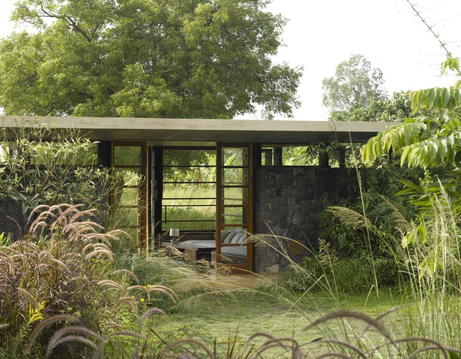 Modern pavilion with dark stone walls and large windows surrounded by dry grasses and green trees.
