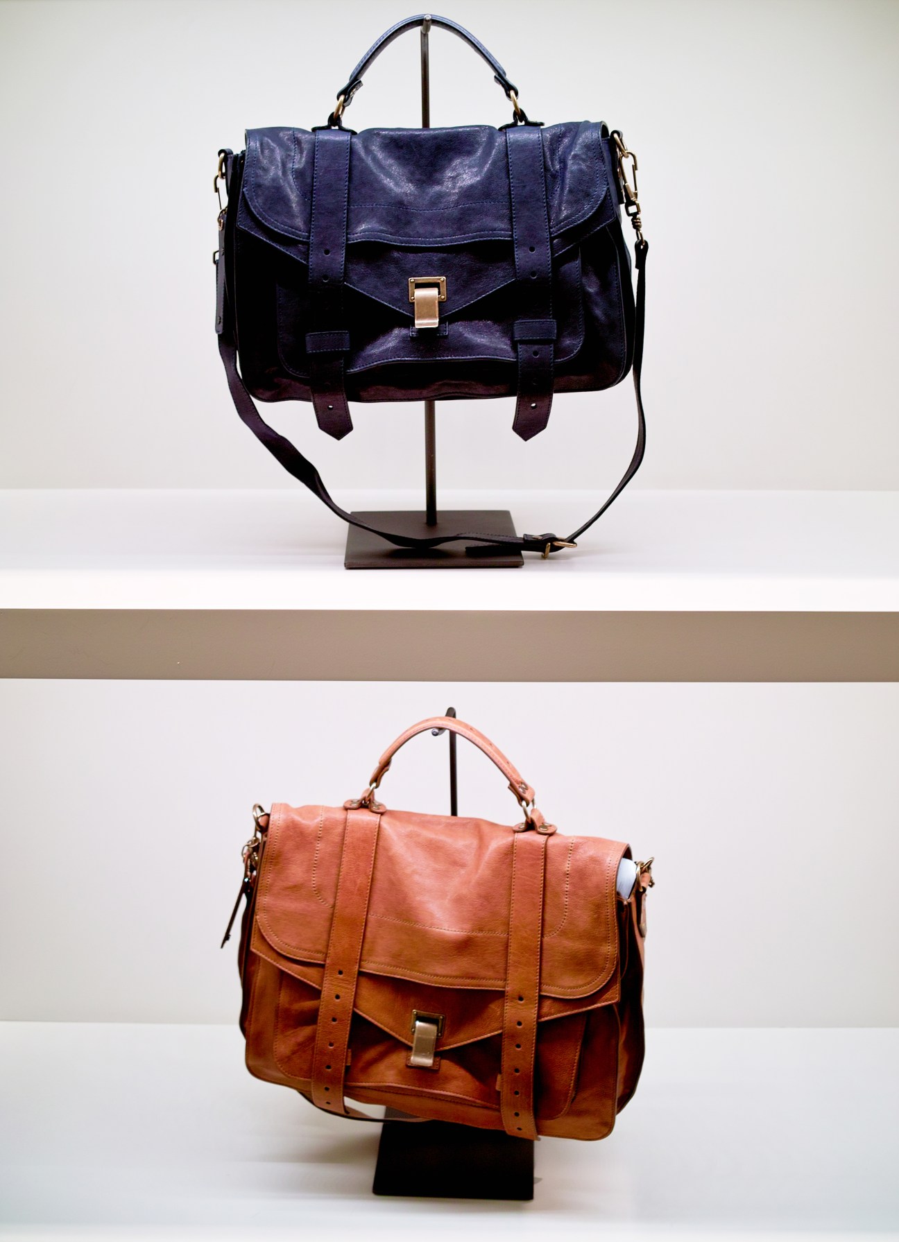 Two Proenza Schouler leather handbags displayed on stands—one navy blue and one cognac brown.