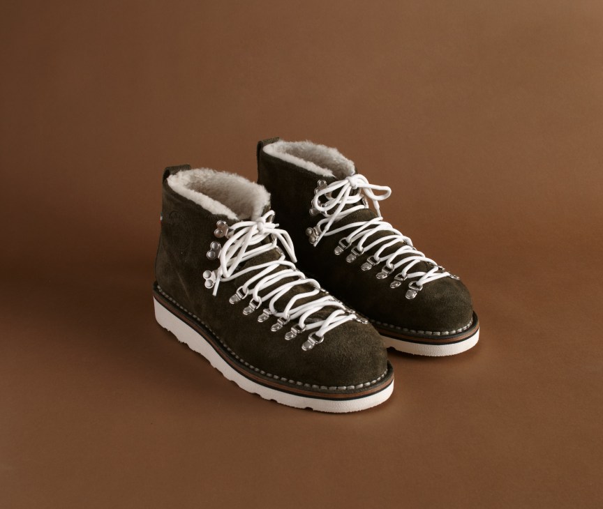 Pair of dark grey suede hiking boots with white laces, sheepskin lining and white rubber soles against a brown background.