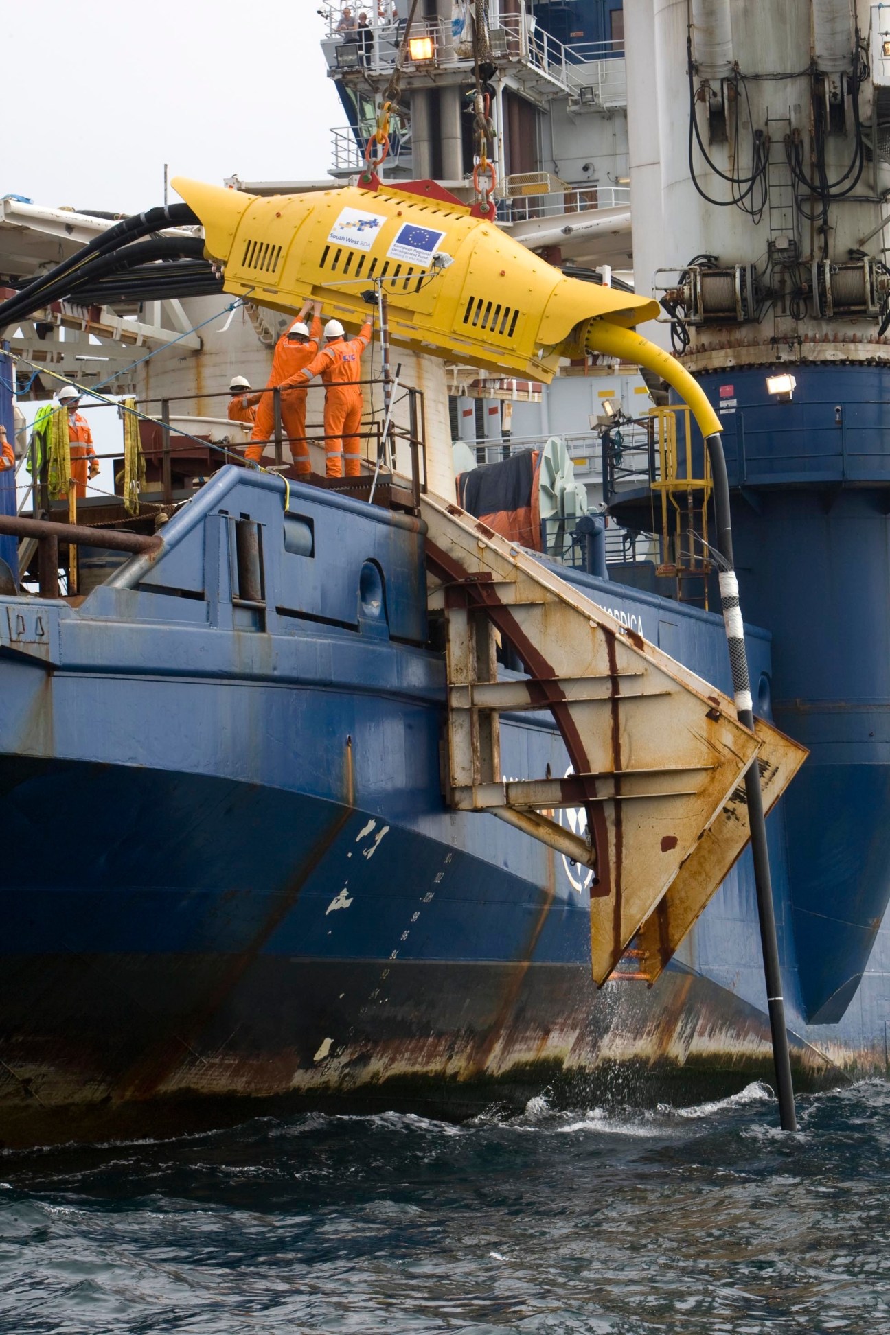 Yellow wave-energy testing device suspended from research vessel with workers in safety gear aboard.